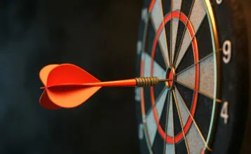 An orange dart hitting the exact center of a bullseye on a dartboard, symbolizing precision, target achievement, and accuracy. This conceptual visual highlights the targeted focus required for students enrolled in the top Chemistry classes in Dwarka, Gurugram & Delhi NCR. The close-up shot captures the dart perfectly embedded in the red inner ring against a moody, dark background, serving as a metaphor for hitting academic goals in competitive exams. Designed as a motivational graphic for expert chemistry classes for JEE & NEET in Dwarka, Janakpuri, Gurugram & Delhi NCR, it reflects the meticulous preparation style offered by Premium Chemistry classes in Janakpuri, Gurugram and Delhi NCR.